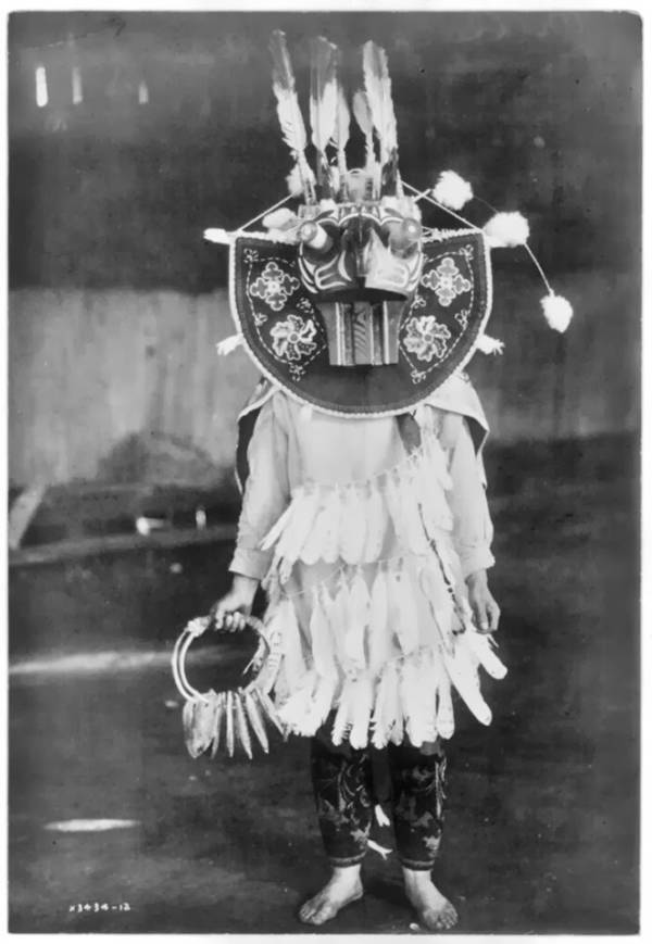 Χορεύτρια με υπερμεγέθη μάσκα, τρεις κρίκους από φτερά μπροστά από τα ρούχα, κρατώντας μια κουδουνίστρα, 1913. (πηγή: Edward Curtis/Library of Congress)